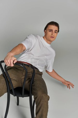 schöner stylischer Mann in Debonair-Kleidung sitzt auf einem Stuhl und schaut in die Kamera vor grauem Hintergrund