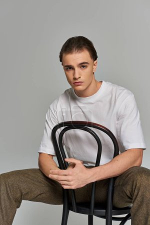 schöner trendiger Mann in Debonair-Kleidung sitzt auf einem Stuhl und blickt in die Kamera vor grauem Hintergrund