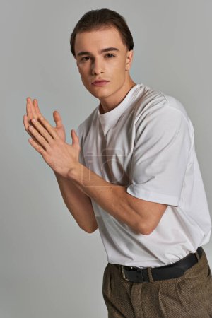 fashionable young male model in sophisticated pants looking at camera while on gray background