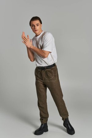 modelo masculino joven de moda en pantalones sofisticados mirando a la cámara mientras está sobre fondo gris