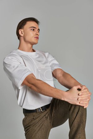 appealing young man in t shirt and brown pants posing attractively on gray backdrop and looking away