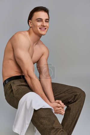 cheerful alluring man in brown pants posing topless while sitting looking at camera on gray backdrop