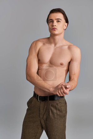 seductor hombre sin camisa en pantalones elegantes marrones posando atractivamente sobre fondo gris y mirando hacia otro lado