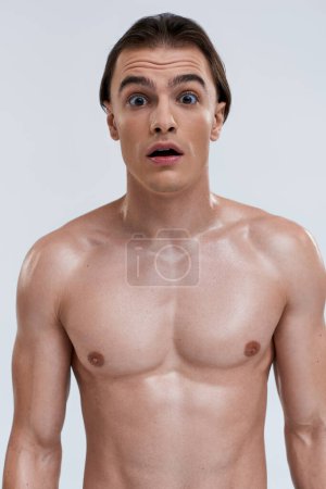 good looking shirtless surprised man posing emotionally on gray backdrop and looking at camera