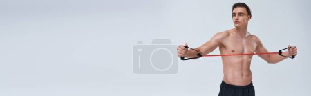 Foto de Tentador hombre deportivo sin camisa en pantalones negros de entrenamiento con expansor de fitness y mirando hacia otro lado, pancarta - Imagen libre de derechos