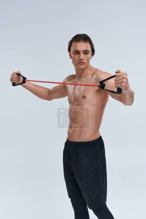 appealing shirtless sporty young man in black pants training with fitness expander and looking away