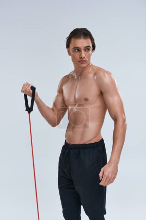 appealing shirtless sporty young man in black pants training with fitness expander and looking away