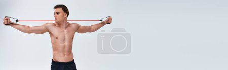 Foto de Atractivo hombre deportivo sin camisa en pantalones negros entrenamiento con expansor de fitness y mirando hacia otro lado, pancarta - Imagen libre de derechos