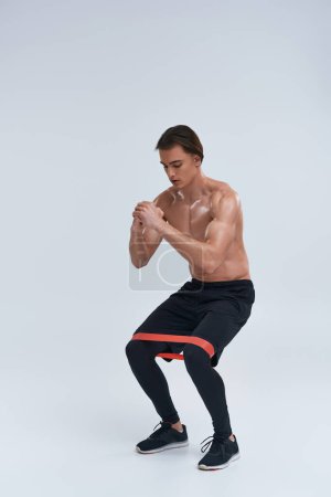 atlético sexy hombre sin camisa en pantalones negros usando banda de resistencia para ponerse en cuclillas sobre fondo gris