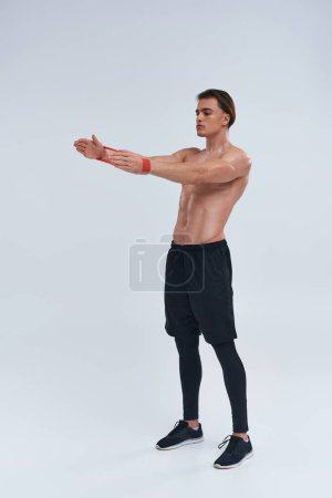 alluring athletic young man in black pants posing topless with resistance band and looking away