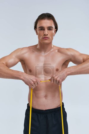 joven atractivo deportivo posando en topless mientras entrena con banda de resistencia y mirando a la cámara