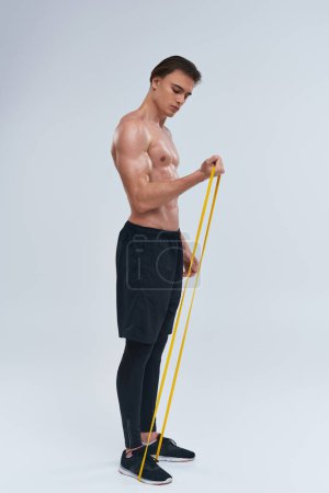 seductor joven deportista sin camisa en pantalones negros ejercitando con banda de resistencia sobre fondo gris