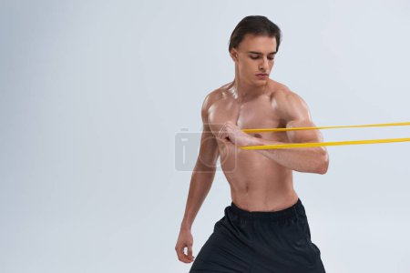 tempting shirtless young sportsman in black pants exercising with resistance band on gray backdrop