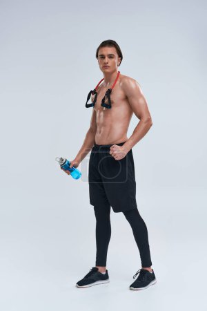 athletic sexy young man posing topless with water bottle and fitness expander and looking at camera