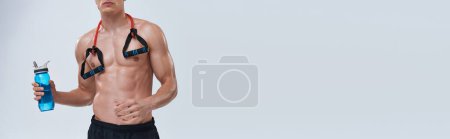 vista recortada de hombre deportivo en pantalones negros posando en topless con botella y expansor de fitness, pancarta