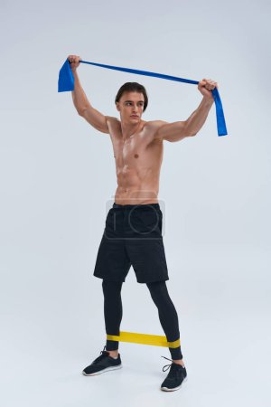 alluring sporty young man in black pants posing topless with resistance bands and looking away