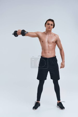 attraktiver athletischer Mann posiert oben ohne beim aktiven Training mit der Hantel und blickt in die Kamera