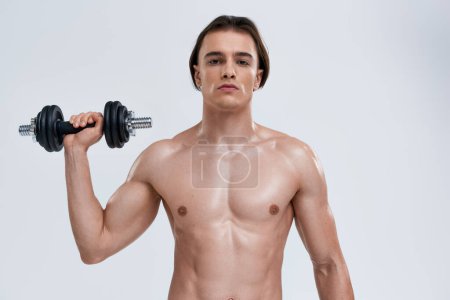 hombre atlético bien parecido posando en topless ejercitando activamente con la mancuerna y mirando a la cámara