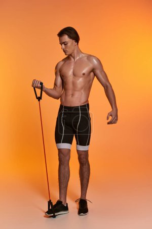 appealing young sporty man in black shorts posing topless and exercising with fitness expander