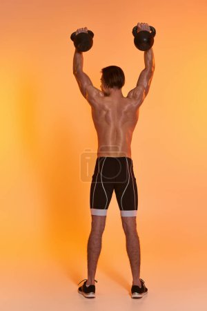 back view of young muscular man posing topless while exercising with kettlebells on orange backdrop
