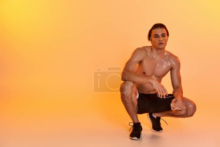 hombre sexy sin camisa en pantalones cortos negros haciendo ejercicio activamente y mirando a la cámara en el fondo naranja
