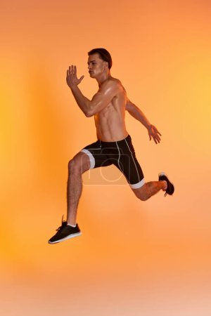attractive shirtless man in black shorts exercising actively and looking away on orange backdrop