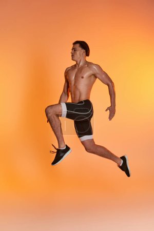 appealing shirtless man in black shorts exercising actively and looking away on orange backdrop