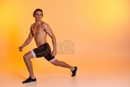 atractivo hombre sin camisa en pantalones cortos negros haciendo ejercicio activamente y mirando hacia otro lado en el fondo naranja