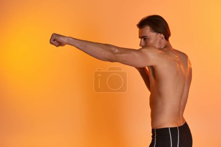 Photo for Alluring shirtless sexy man in black sporty shorts boxing actively on vibrant orange background - Royalty Free Image