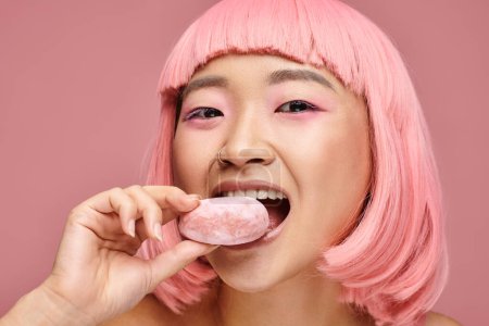 retrato de feliz asiático joven mujer con rosa pelo comer mochi contra vibrante fondo