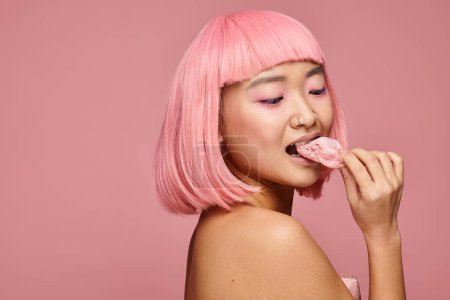 Charmante asiatische Frau in ihren Zwanzigern mit pinkfarbenen Haaren, die Mochi über der Schulter vor lebendigem Hintergrund essen