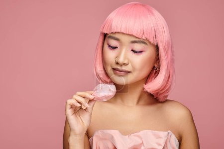 süße asiatische Frau in ihren Zwanzigern mit rosa Haaren und Make-up, die auf Mochi vor lebendigem Hintergrund schaut