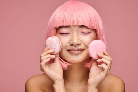 happy asian woman with pink hair and closed eyes posing with sweets in vibrant background