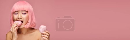 horizontal view of charming asian woman in 20s with pink hair eating sweets on vibrant background