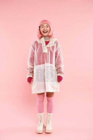 happy and pretty asian woman with pink hair in rain coat posing against vibrant background