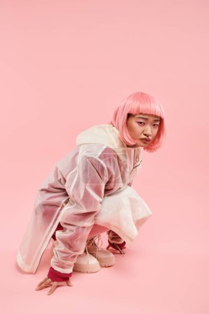 lindo asiático mujer en 20s con rosa pelo en elegante traje agachado abajo contra vibrante fondo