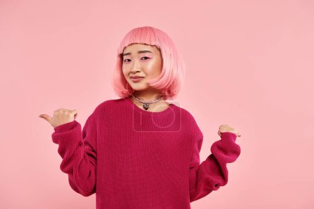 mujer joven en vibrante jersey y pelo rosa apuntando con sus pulgares y mirando a la cámara