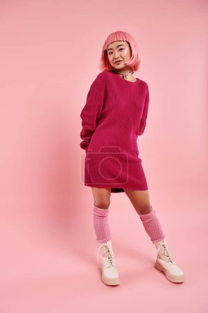 playful young woman in vibrant sweater dress with pearl necklace posing against pink background