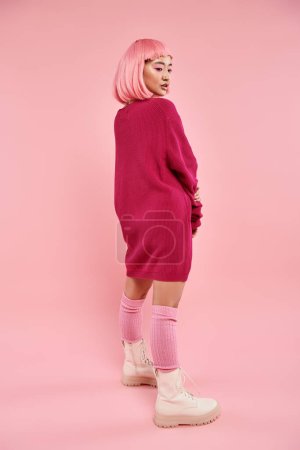 pretty asian woman in vibrant sweater looking to down over shoulder in pink background