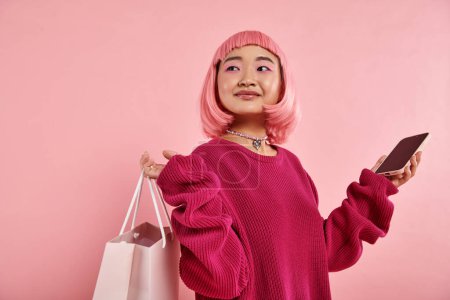 Téléchargez les photos : Jolie asiatique jeune femme avec shopping paquet et smartphone cherche à côté sur fond rose - en image libre de droit