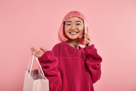 Téléchargez les photos : Jeune femme souriante appelant du smartphone et tenant le paquet sur l'épaule sur fond rose - en image libre de droit