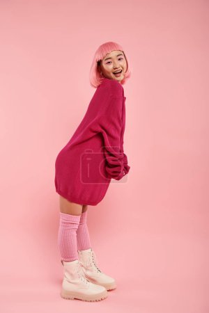 fröhliche Asiatin im großen Pullover-Outfit steht in seitlicher Pose vor rosa Hintergrund