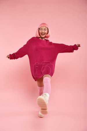 Photo for Happy smiling asian young woman in stylish outfit posing with leg raised against pink background - Royalty Free Image