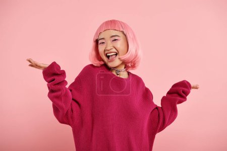 alegre asiático joven mujer en Grande suéter shrugging su hombros felizmente en rosa fondo