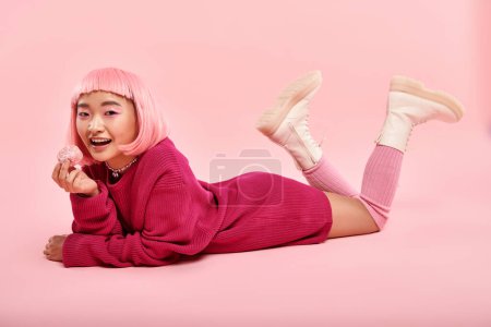juguetona mujer asiática en vibrante traje de suéter acostado con mochi sobre fondo rosa