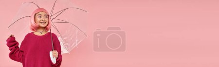 Photo for Horizontal shot of cute asian woman standing under umbrella and looking to side on pink background - Royalty Free Image