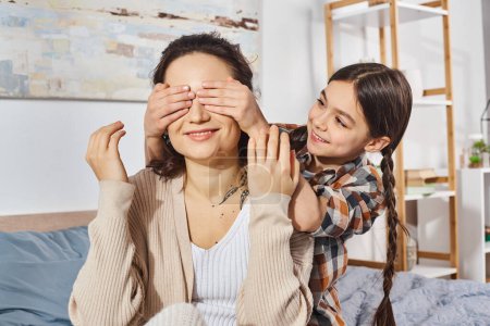 A girl covering eyes of her mother, sharing a playful moment together at home.