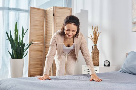 A woman stands gracefully over a bed in a bedroom, making blanket wrinkles