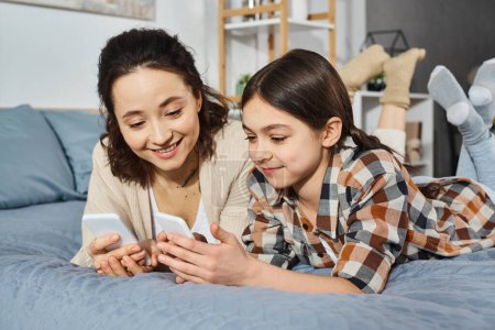 Mutter und Tochter liegen auf dem Bett, konzentrieren sich auf den gemeinsamen Handy-Bildschirm, genießen die gemeinsame Zeit.