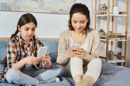 mother and daughter, sitting on a bed, focused on their phones, sharing a moment of digital connection.